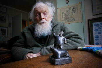 Photograph of Adam Watson later in life.  On a table in front of him is a stone and ivory carving of a hunter and seal